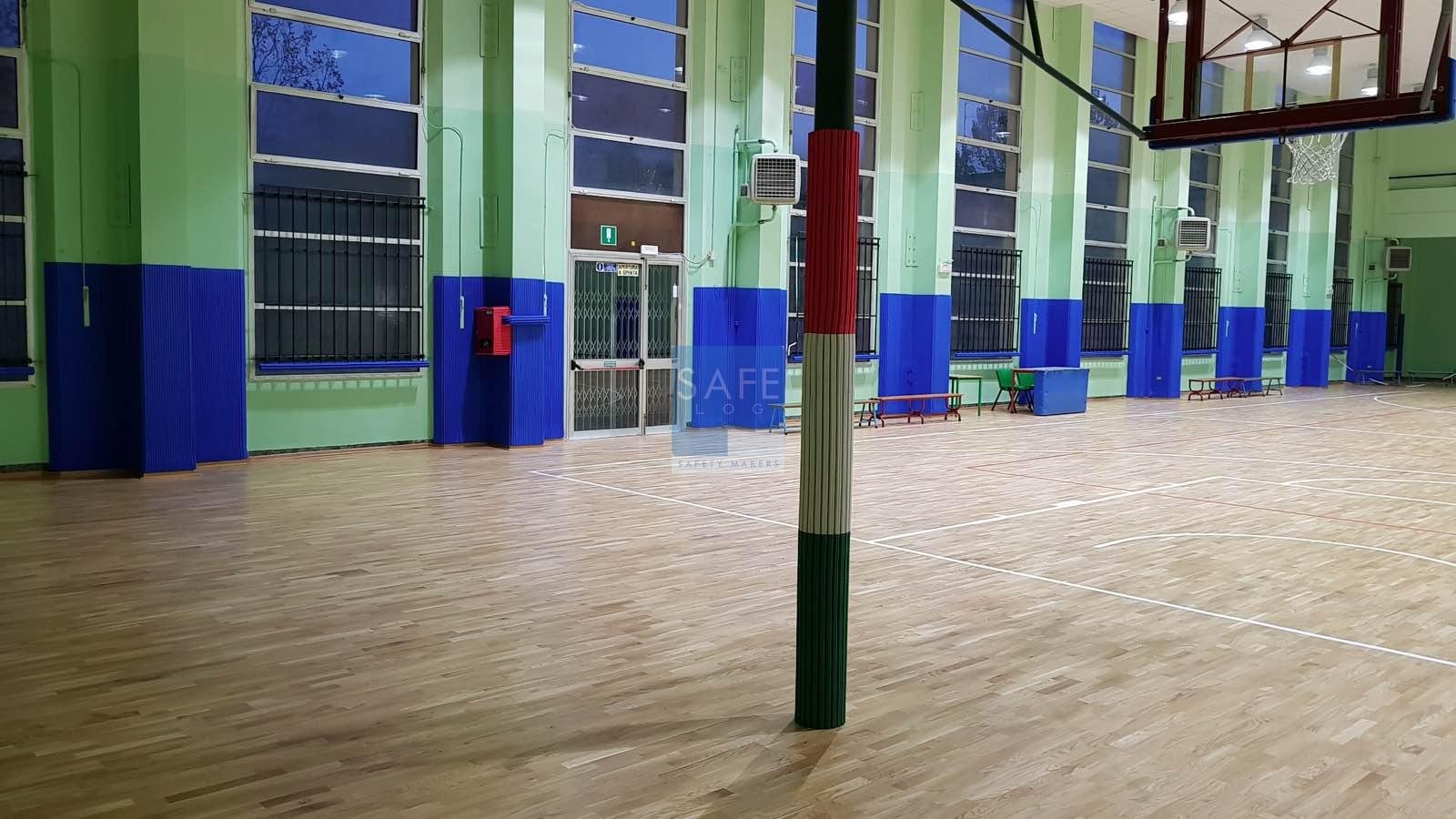 Pilastri e sporgenze di palestra scolastica rivestiti con protezioni antitrauma blu certificate, con palo tabellone basket decorato con colori tricolore italiani