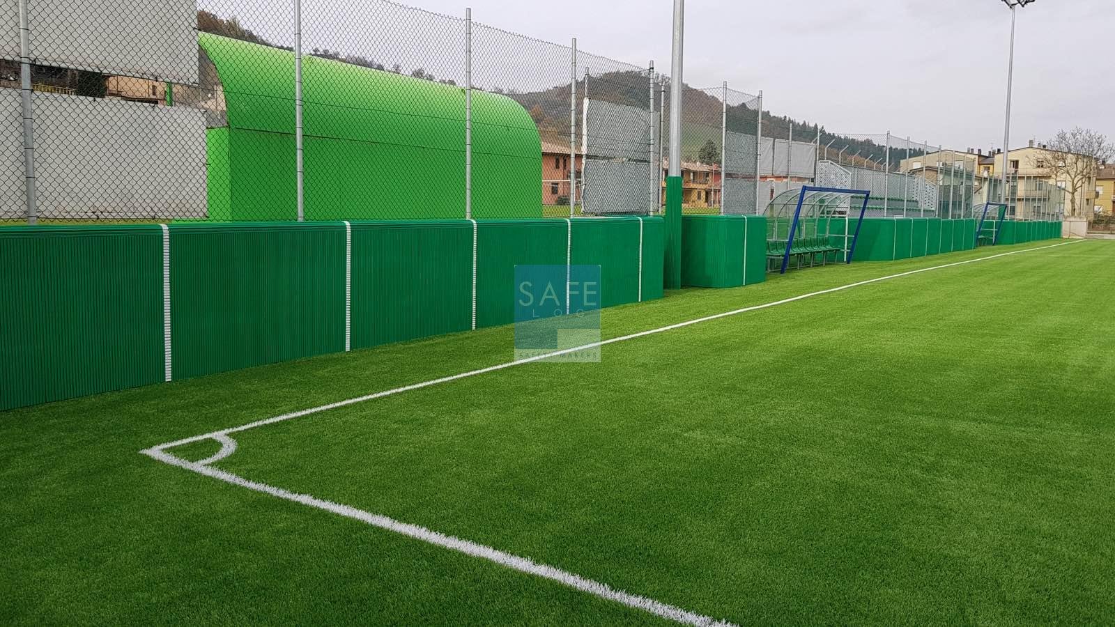 Protezioni antitrauma in gomma verde installate su pali, panchine e muretti a bordo campo in un campo da calcio in erba sintetica ad Amatrice per garantire la sicurezza degli atleti.